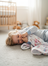 Load image into Gallery viewer, Child snuggled with blanket, lying on a carpeted floor in a room with a crib and toys.